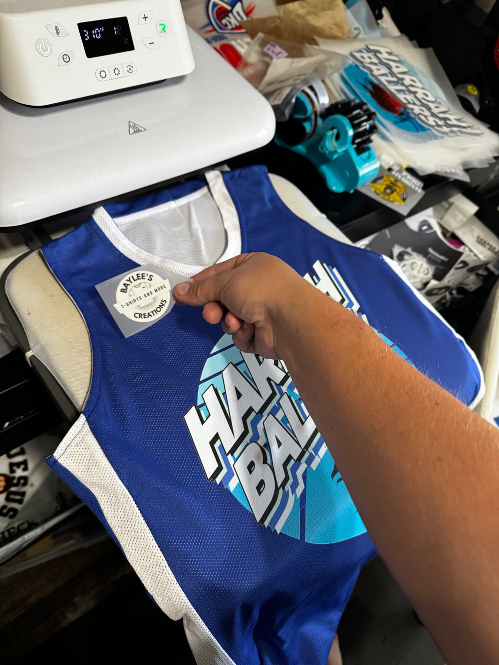 A blue and white basketball jersey with a custom graphic print, being held by a hand near a heat press machine.
