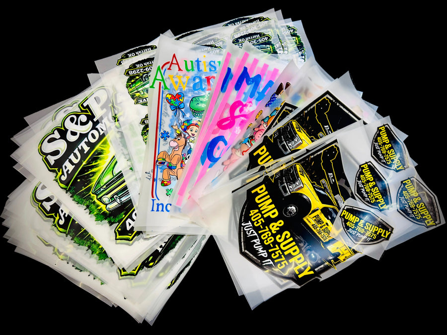 A pile of DTF Prints, including designs with 'S&P CAUTION', 'Auto Parts Warehouse', 'PUMP & SUPPLY', and cartoon graphics, arranged on a black textured background.